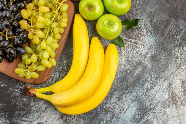 상위 클로즈업 보기 과일 3개의 바나나 사과 녹색 및 검은색 포도가 부엌 보드에 있습니다.