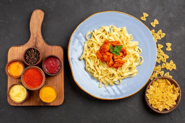 暗いテーブルのまな板のパスタとスパイスとソースのボウルの横にある肉汁と肉の食欲をそそるパスタのパスタの青いプレートの上部の拡大図プレート