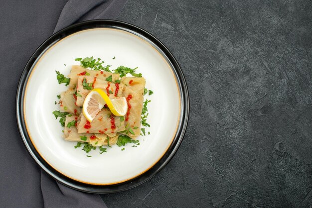 Top close-up view plate on tablecloth white plate of stuffed cabbage with herbs lemon and sauce on grey tablecloth on the left side of dark table