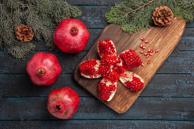 Top close-up view pomegranate and branches pilled pomegranate on wooden board next to seeds of pomegranate spruce branches with cones knife and three pomegranates on grey table