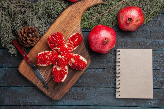 Top close-up view red pomegranate on board pilled pomegranate on cutting board next to ripe three pomegranates knife white notebook and spruce branches and cones on table