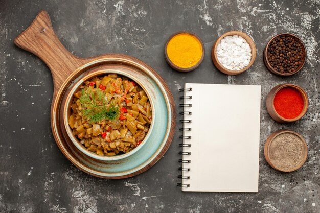 Top close-up view spices and dish bowls of different spices next to the white notebook and dish of green beans and tomatoes on the wooden tray on the black table