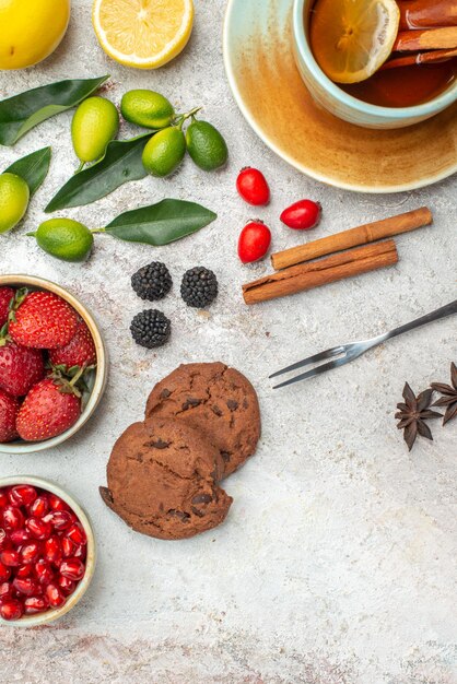 Top close-up view strawberries cookies berries in bowls cookies a cup of tea lime and lemon cinnamon sticks fork on the table