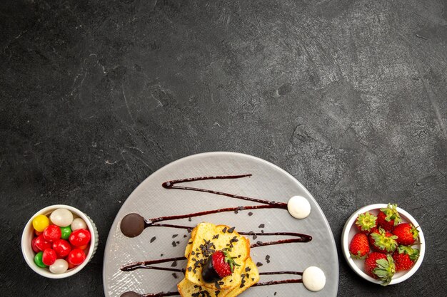 Top close-up view sweets grey plate of pieces of cake with chocolate sauce and strawberries next to the bowls of candies and strawberries