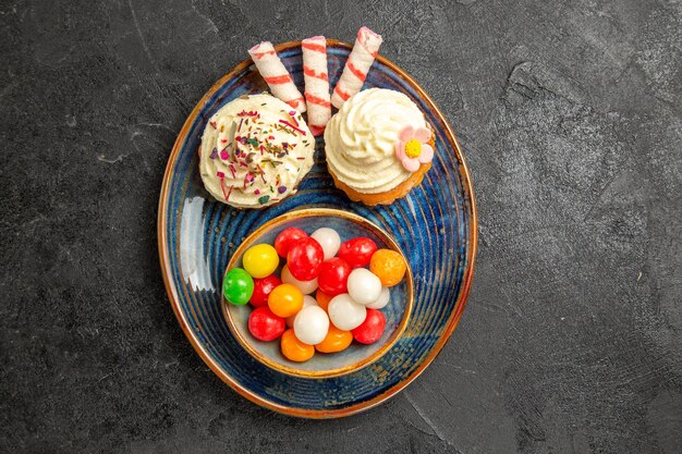 Сверху крупным планом вид сладостей на тарелке аппетитных кексов рядом с миской разноцветных конфет на темном столе