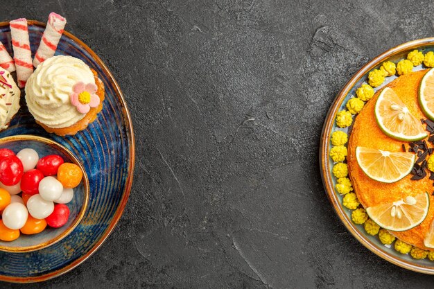 Top close-up view sweets on the plate appetizing cupcakes and bowl of sweets next to the cake with slices of citrus fruits on the black table