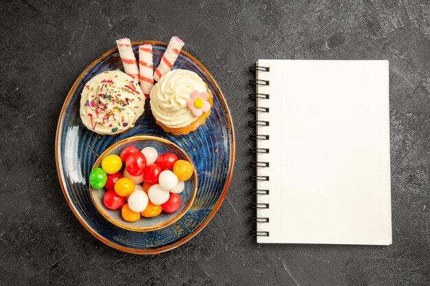 Top close-up view sweets on the plate white notebook next to the blue plate of the appetizing cupcakes and bowl of colorful sweets on the dark table