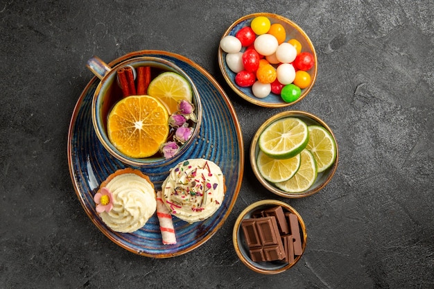 Top close-up view sweets on the table bowls of the appetizing candies chocolate and slices of limes next to the blue saucer of the cup of black herbal tea and two cupcakes on the table