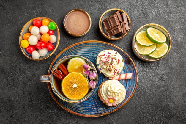 Top close-up view sweets on the table four bowls of chocolate candies and limes a plate of two cupcakes and the cup of herbal tea on the table