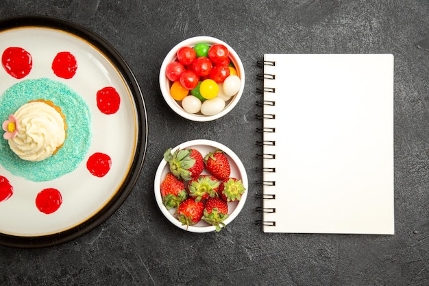 컵케이크 접시 옆에 있는 탁자 흰색 공책에 있는 클로즈업 보기 과자와 검정 탁자에 있는 과자와 딸기 그릇