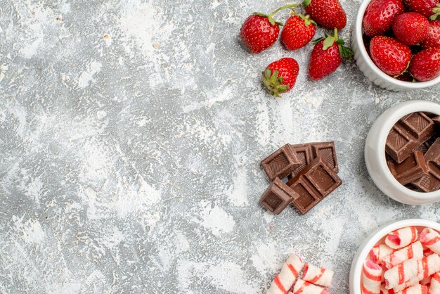 Top close view bowls with strawberries chocolates candies and some strawberries chocolates candies at the right side of the grey-white ground