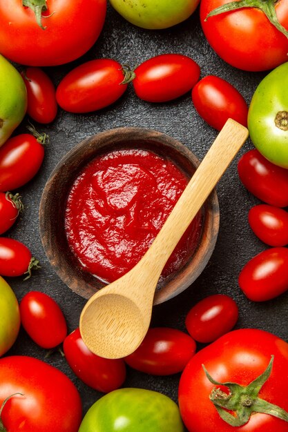 Top close view cherry red and green tomatoes around a bowl with ketchup and a wooden spoon on dark ground