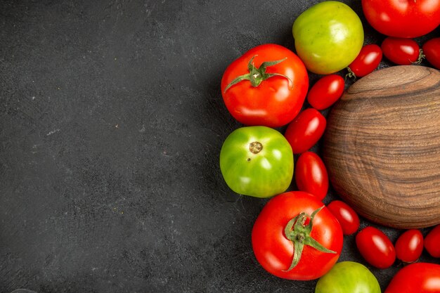 Top close view cherry red and green tomatoes around a wooden plate on dark table with free space