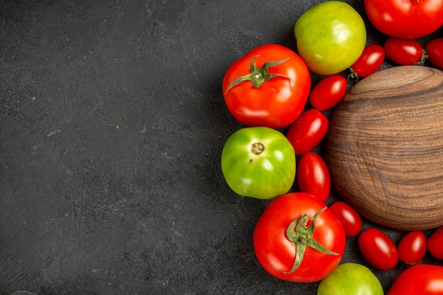 Top close view cherry red and green tomatoes around a wooden plate on dark table with free space