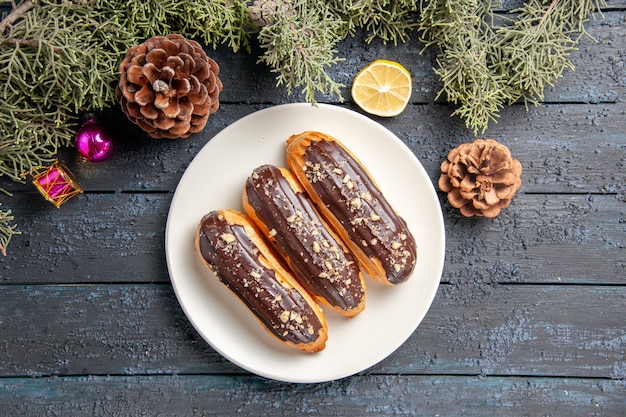 흰색 타원형 접시 콘 전나무 나무에 상위 닫기보기 초콜릿 eclairs 복사 공간이 어두운 나무 바닥에 레몬의 크리스마스 장난감 조각을 나뭇잎