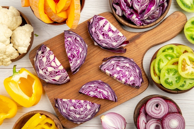 Top close view chopped red cabbage on wood board cut pupmkin cut green tomatoes cut yellow bell peppers in bowls on white surface