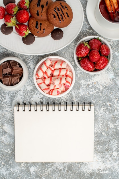 Foto gratuita vista ravvicinata dall'alto biscotti fragole e cioccolatini rotondi sul piatto ovale ciotole di caramelle fragole cioccolatini tè alla cannella e un taccuino sul tavolo grigio-bianco