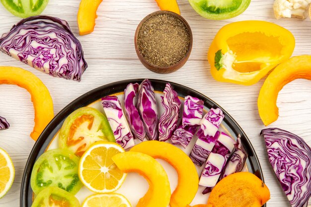 Top close view cut vegetables and fruits pumpkin persimmon red cabbage lemon green tomatoes yellow bell peppers on black platter black pepper in bowl on table