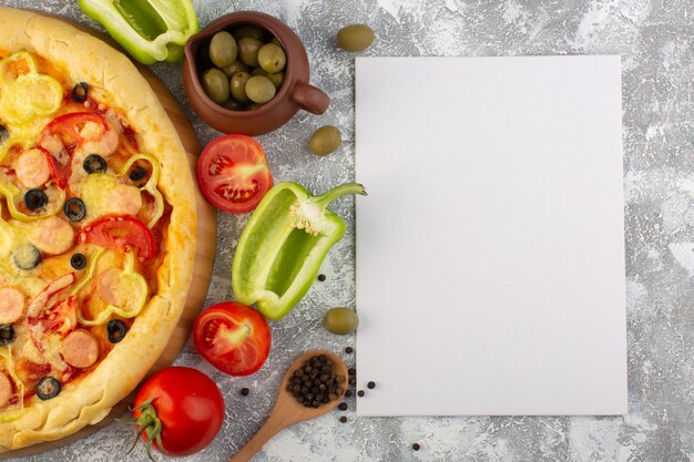 Вид сверху вкусной сырной пиццы с оливками, сосисками и помидорами на сером столе с бумажной пустой едой из итальянского теста быстрого питания