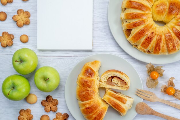 Top close view of delicious sliced pastries inside plate with filling along with green apples notepad and cookies on white desk, pastry cookie biscuit sweet