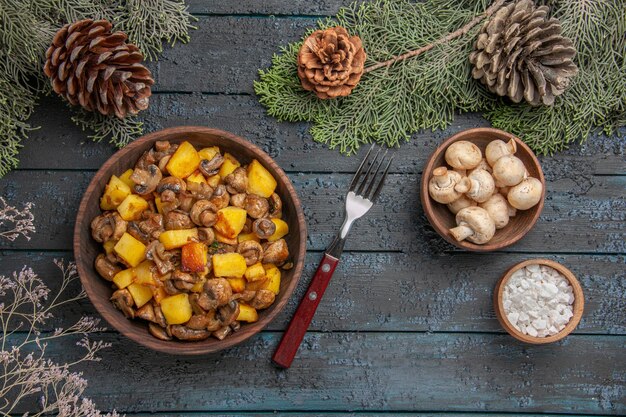 가문비나무 가지 아래 회색 테이블에 있는 상단 닫기 접시와 버섯과 감자의 가지 접시와 포크 버섯과 소금 옆에 원뿔이 있습니다.
