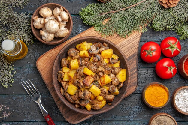 Top close view dish and vegetables dish of potatoes and mushrooms on board next to fork three tomatoes and colorful spices under oil in bottle tree branches and bowl of mushrooms