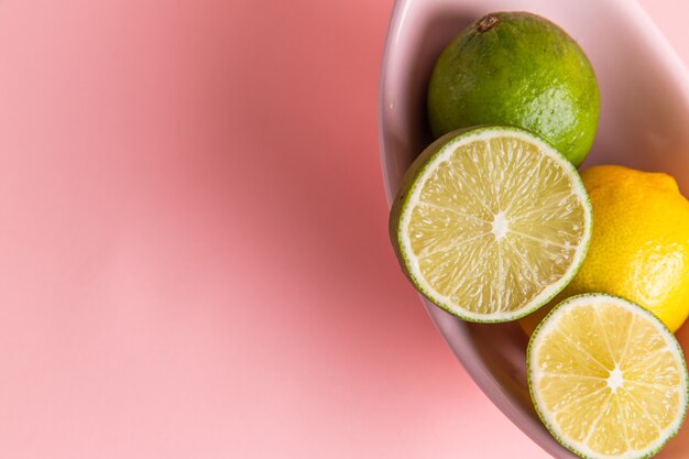 Top close view of fresh lemons with sliced lime inside plate on light-pink surface