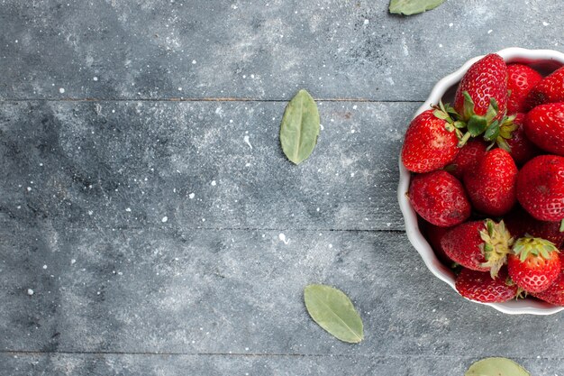 Top close view of fresh red strawberries inside white plate along with green dried leaves on grey wooden , fruit fresh berry color photo vitamine health