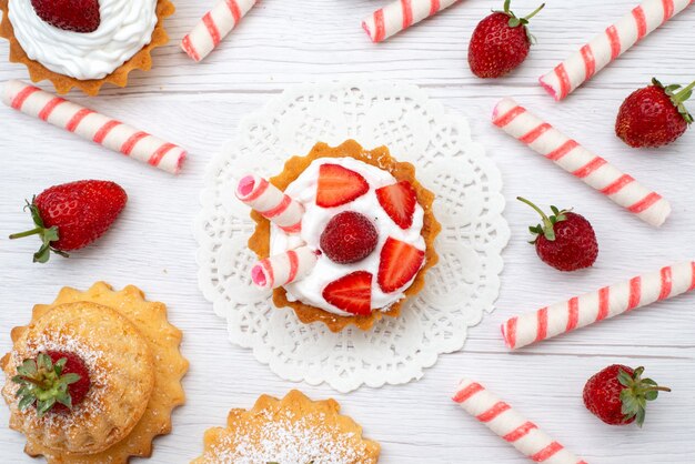 Top close view of little yummy cakes with cream and sliced strawberries candies on white