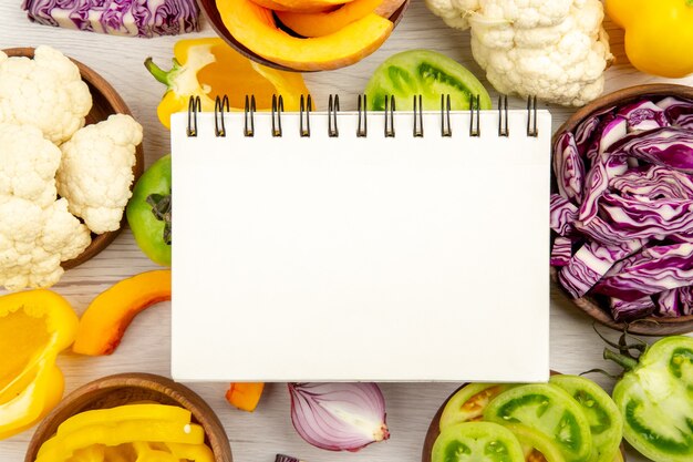 Top close view notepad cut green tomatoes cut red cabbage cut pumpkin cauliflower cut bell peppers in bowls on surface