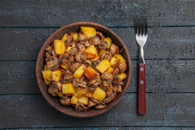 Top close view plate and fork bowl with appetizing potatoes and mushrooms next to the fork on dark table