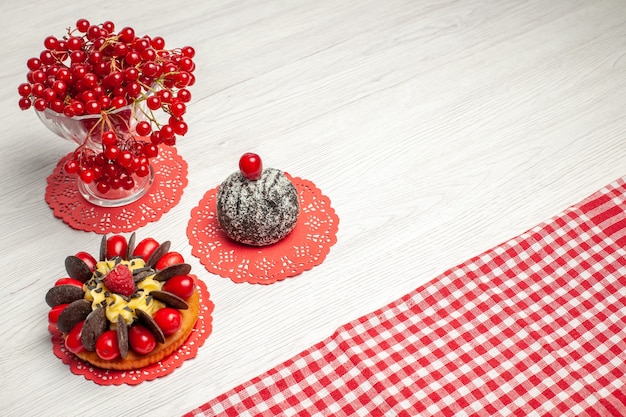 Сверху крупным планом красная смородина в хрустальном ягодном пироге и какао-пирог на красной овальной кружевной салфетке и красно-белая клетчатая скатерть на белом деревянном столе