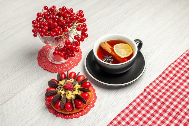 Top close view red currant in a crystal glass on the red oval lace doily and a cup of lemon cinnamon tea and red-white checkered tablecloth on the white wooden table