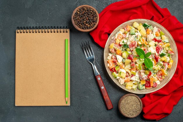 Top close view of veggie salad on red napkin with fork notepad and pepper on side on dark grey background