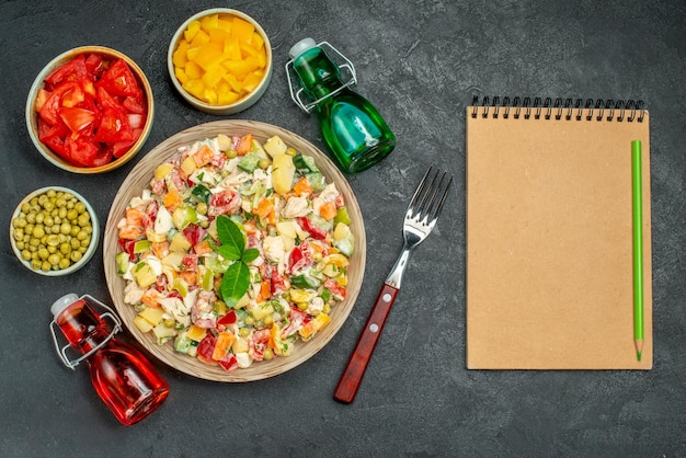 Top close view of veggie salad with fork bottles of oil and vinegar bowls of veggies and notepad on side on dark grey table