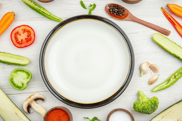 Top close view white round plate cut vegetables spices in bowls wooden spoon on white background