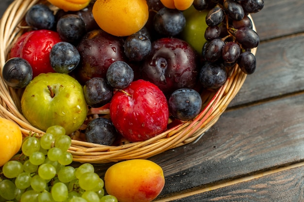Корзина с фруктовыми спелыми и кислыми фруктами, такими как виноград, абрикосы, сливы, на коричневом деревенском столе.