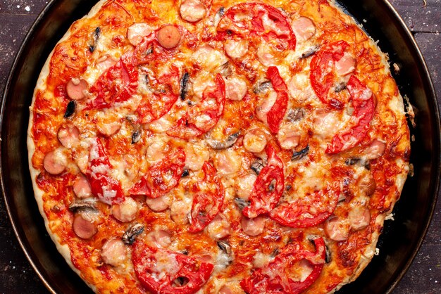 top closer view of cheesy tomato pizza with olives and sausages inside pan on brown desk