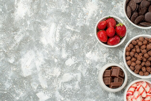 Top distant view bowls with candies strawberries bitter and milky chocolates cereals and cacao at the right side of the grey-white ground