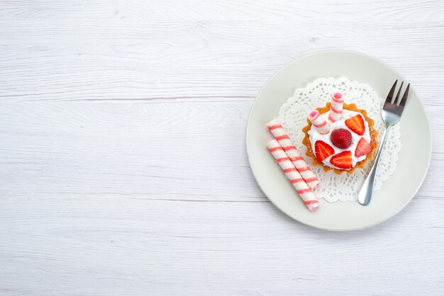 Вид сверху на маленький торт со сливками и нарезанной клубникой внутри тарелки на белом, фруктовый торт, ягодный сладкий сахар