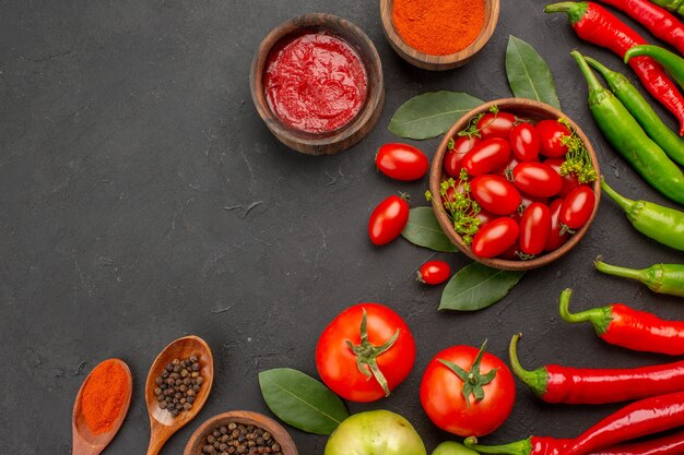 Top half view a bowl of cherry tomatoes hot red and green peppers bay leaves spices in wooden spoons bowls of ketchup hot red pepper powder and black pepper and tomato on ground