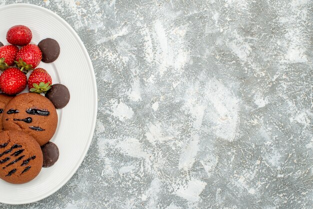 Top half view chocolate cookies strawberries and round chocolates on the white oval plate at the left side of the grey-white ground