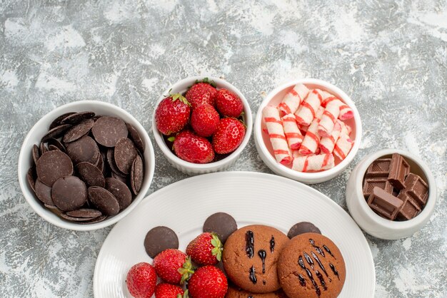 Верхняя половина вида печенье с клубникой и круглые шоколадные конфеты на белой овальной тарелке, окруженной мисками с конфетами, клубникой и шоколадными конфетами на земле