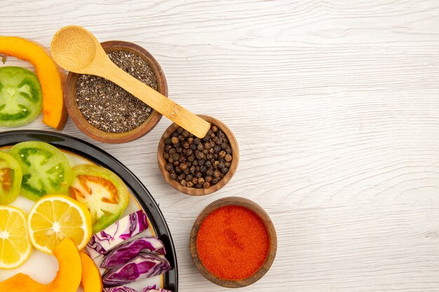 Top half view cut vegetables and fruits pumpkin bell peppers persimmon red cabbage on black plate spice in small bowls wooden spoon on wooden table copy space