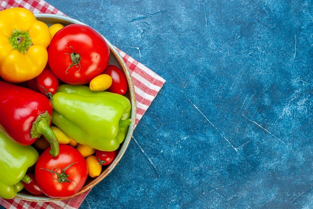 Top half view fresh vegetables cherry tomatoes different colors bell peppers tomatoes cumcuat on platter on red and white checkered tablecloth on blue table with free space