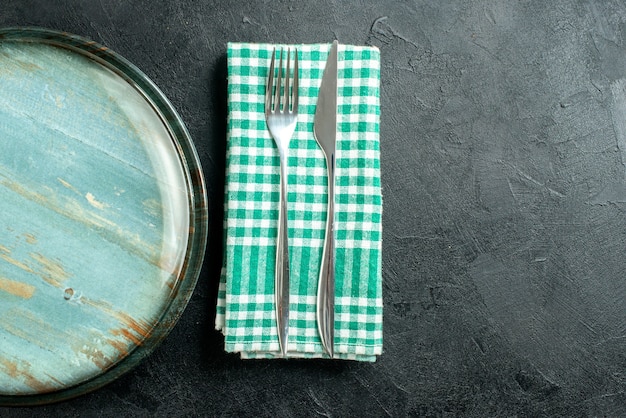 Top half view round platter dinner knife and fork on green and white checkered napkin on black table copy place