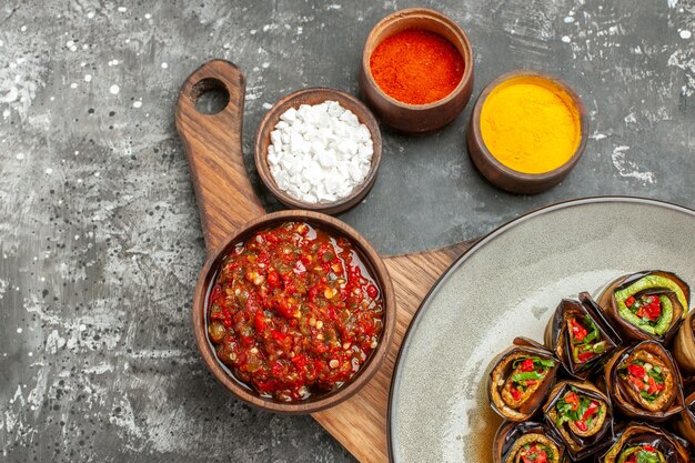 Top half view stuffed aubergine rolls in oval plate adjika in bowl on wooden serving board with handle different spices in small bawls on grey-white background