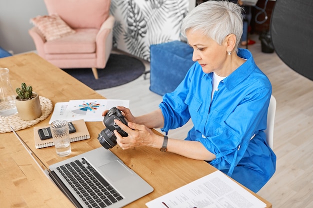 세련된 중년 여성 사진 작가가 열린 노트북으로 직장에 앉아 DSLR 카메라를 들고 수정을 위해 최고의 사진을 선택하고 집중된 표현을 집중시킨 최고 높은 각도보기