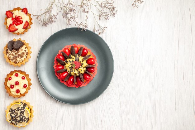 Top left side view berry cake in a grey plate and from top to the bottom different tarts on the white wooden ground