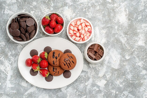 Top left side view cookies strawberries and round chocolates on the white oval plate surrounded bowls with candies strawberries and chocolates at the left side of the ground